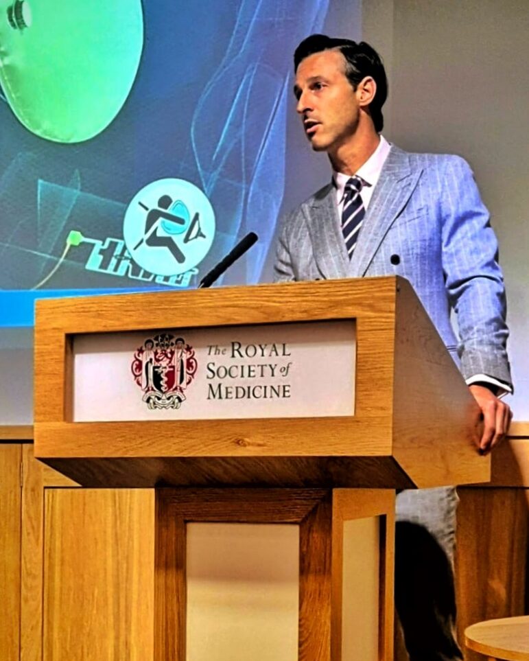 a surgeon in a suit standing at a lectern labelled with the Royal Society of Medicine, delivering a lecture